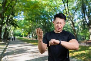 dérangé Jeune homme asiatique athlète permanent dans parc et à la recherche à intelligent montre, vérification résultat, impulsion, temps. il se répand le sien mains insatisfait. photo