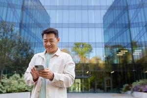 réussi asiatique homme dans décontractée vêtements les usages le téléphone, homme des promenades dans le ville près une moderne bâtiment, cadrans une message, appels, homme d'affaire après travail. photo