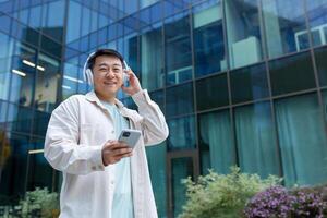 portrait de asiatique homme en plein air avec téléphone et écouteurs pour écoute à musique, content homme en utilisant téléphone intelligent app souriant et à la recherche à caméra. photo