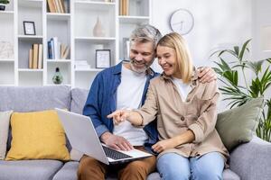 Sénior mature couple homme et femme séance à Accueil dans vivant pièce sur canapé, gens dans l'amour en utilisant ordinateur portable, ayant amusement choisir des produits dans en ligne l'Internet boutique ensemble. photo