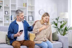 excité effrayé homme est appel un ambulance sur le téléphone car le sien épouse est malade, elle est en portant sa cœur avec sa main, elle est respiration difficile de douleur à maison, cœur attaque. photo