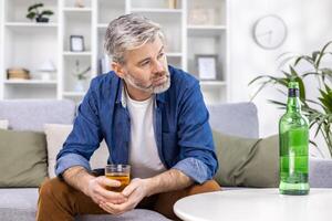 proche en haut de adulte mature homme solitaire déprimé en buvant fort de l'alcool séance sur canapé dans vivant pièce sur journée. photo