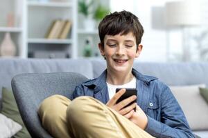 portrait de une souriant adolescent garçon enfant séance sur le canapé à Accueil et en utilisant le téléphone. bavardage, est assis dans social les réseaux, pièces en ligne Jeux. photo