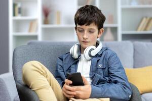 sérieux enfant de une adolescent garçon dans écouteurs séance à Accueil sur le canapé, en utilisant le téléphone. écoute à musique, montres vidéos. il chats, est assis sur social les réseaux, pièces en ligne Jeux. photo