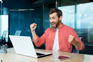 une Jeune homme est assis dans le bureau, regards à le portable moniteur et se réjouit. cris avec bonheur, fait du victorieux main gestes. reçu bien nouvelles, réussi accord, projet, en jouant en ligne Jeux. photo