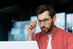 fermer photo. portrait de une Jeune Masculin programmeur, une homme d'affaire qui est se concentrer sur travail dans le Bureau à une ordinateur portable, à la recherche à le moniteur, en portant des lunettes sur le sien affronter. photo