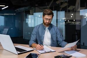 une sérieux Jeune homme comptable, financier, analyste, Auditeur est assis dans le Bureau à le tableau. travaux avec documents. écrit et calcule le budget, finances. photo