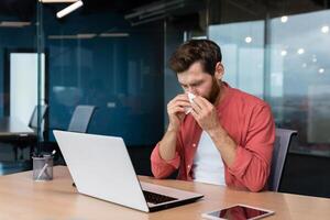 une Jeune homme est malade à travail. est assis à le bureau dans le bureau, lingettes le sien nez avec une serviette de table, a une qui coule nez, Est-ce que ne pas ressentir Bien. photo