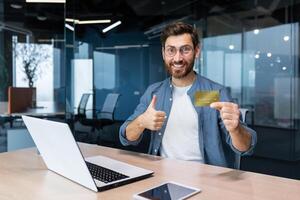 banque crédit carte publicité. une Jeune homme est assis dans le Bureau à le table et détient une d'or argent carte, des offres à acheter, spectacles une super main à le caméra. photo