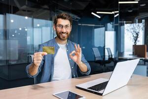 réussi homme d'affaire à l'intérieur Bureau bâtiment à lieu de travail souriant et à la recherche à caméra, mature adulte homme en portant et montrant banque crédit carte, souriant avec contentement. photo