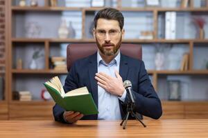 Sénior mature pasteur en train de lire Bible livre en ligne, homme à la recherche à caméra séance dans Accueil Bureau et enregistrement l'audio livre en ligne. photo