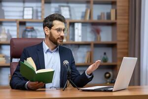 une Jeune homme pasteur de une église, communauté est assis dans le Bureau avec une portable et une microphone et lit une sermon. prières en ligne de le Bible. adresses croyants et paroissiens via appel. photo