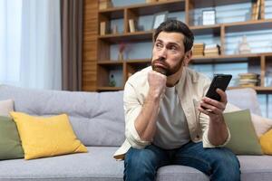 un dérangé Jeune homme est séance sur le canapé à Accueil avec une téléphone dans le sien mains, à la recherche pensivement à le côté. attendre et reçu une message de une petite amie, amoureux, ex, épouse, photo