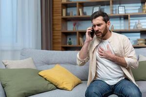 homme ayant cœur attaque à maison, patient appel médecin en utilisant téléphone, séance sur canapé dans vivant pièce à maison. photo