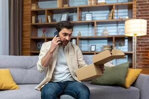 un en colère Jeune homme est séance à Accueil sur le canapé, en portant un ouvert boîte dans le sien mains. je reçu une faux, vide, endommagé colis. pourparlers sur le téléphone avec le livraison service, le magasin. photo