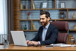 réussi mature homme d'affaire à travail avec portable à l'intérieur bureau, homme dans affaires costume séance à bureau dactylographie sur ordinateur, satisfait avec réussite résultat et travail. photo