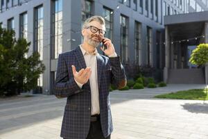 mature homme d'affaire dans élégant costume ayant une animé conversation sur le sien mobile téléphone dans de face de un Bureau bâtiment. photo