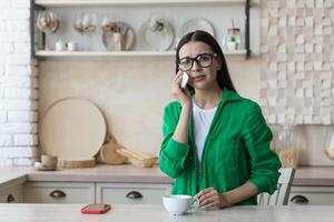 dérangé Jeune femme séance à Accueil dans le cuisine avec une mobile téléphone et pleurs. il regards Malheureusement à le caméra, lingettes le sien larmes avec une serviette de table. photo