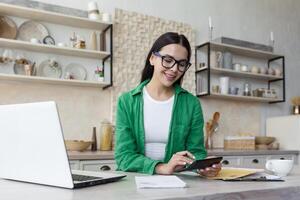 une Jeune fatigué femme est séance à le table dans le cuisine à maison. il travaux sur une portable et avec documents, chèques comptes, calcule le famille budget. affaires revenu. photo