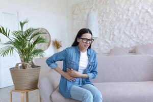 une Jeune femme est séance sur le canapé à maison, grimaçant et en portant sa estomac. souffre de sévère abdominal douleur, menstruel douleur, grossesse problèmes, fausse-couche, empoisonnement, spasmes. photo