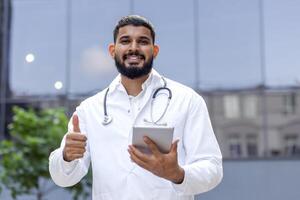 portrait de une souriant Jeune Indien Masculin médecin, une étudiant permanent près une clinique Université dans une blanc manteau, en portant une tablette et montrer du doigt à le super caméra. photo