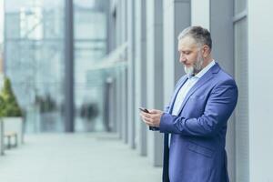 sérieux et sur de soi aux cheveux gris homme permanent à l'extérieur Bureau bâtiment, homme d'affaire Sénior banquier dans affaires costume en portant téléphone, réussi investisseur en utilisant téléphone intelligent photo