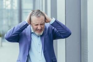 Sénior aux cheveux gris homme d'affaire à l'extérieur Bureau bâtiment déprimé, homme dans désespoir et abattement, faillite investisseur perdu argent, en portant mains sur tête photo