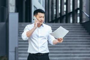 frustré Assurance agent avec les documents parlant sur le téléphone à l'extérieur le bureau, déprimé asiatique homme d'affaire photo