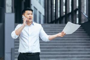 frustré Assurance agent avec les documents parlant sur le téléphone à l'extérieur le bureau, déprimé asiatique homme d'affaire photo