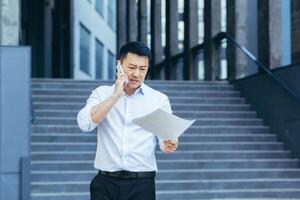 frustré et en colère asiatique homme d'affaire parlant sur le téléphone, en portant une lettre et les documents de une banque à l'extérieur le Bureau photo