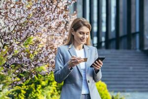 magnifique blond femme près une arbre avec fleurs détient une téléphone et une banque crédit carte, sourit et fait du achats dans le en ligne boutique photo