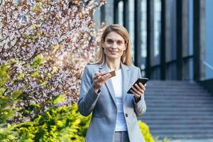 content affaires femme près Bureau bâtiment regards à caméra et sourires, les usages téléphone et banque crédit carte pour achats dans en ligne boutique photo