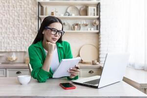 une Jeune femme d'affaires dans des lunettes est assis à Accueil dans le cuisine avec une portable et conduit un en ligne éloigné entretien pour le emploi de Nouveau employés. écoute soigneusement, détient une carnet de notes, prend Remarques. photo