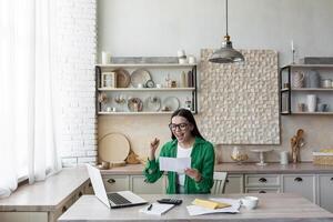 content Jeune fille étudiant dans des lunettes et vert chemise se réjouit, reçu lettre de invitation à université. reçu notification de admission. il est assis à le table à maison, ouvre un enveloppe. photo