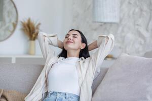 portrait content Jeune magnifique femme relaxant à Accueil sur canapé avec yeux fermé, bras derrière diriger. jouit silence, paix, rêves, sourit. avoir une agréable fin de semaine. photo