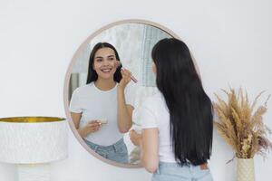 portrait de une Jeune magnifique femme permanent dans de face de une miroir et appliquant maquillage avec une brosse sur sa affronter. Aller à travail, une réunion, une marcher, souriant photo