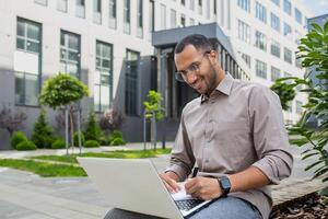 Jeune hispanique étudiant en train d'étudier séance sur banc à l'extérieur Université Campus, homme en utilisant portable à regarder séminaire en ligne, cours, l'écriture Les données dans carnet souriant avec contentement. photo