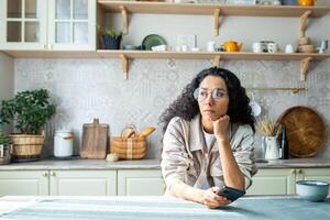 solitaire ennuyé femme séance seul à Accueil dans cuisine, hispanique femme avec frisé cheveux déprimé en pensant en portant téléphone, eu mal nouvelles message. photo