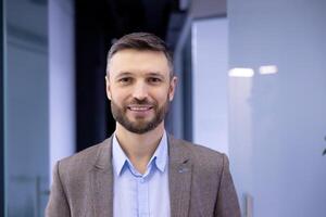 fermer portrait de mature réussi homme d'affaire, homme souriant et à la recherche à caméra à l'intérieur Bureau bâtiment à lieu de travail, satisfait banque investisseur chef. photo