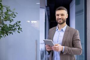 portrait de une souriant Jeune homme d'affaire permanent dans le hall de un Bureau centre, en portant une tablette et à la recherche à le caméra. photo