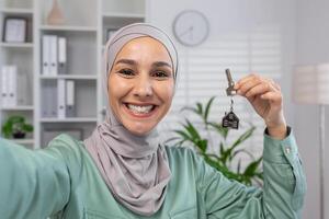 une de bonne humeur femme dans une hijab fièrement montrant une maison clé, symbolisant Nouveau Accueil la possession et une Frais début dans sa confortable intérieur paramètre. photo