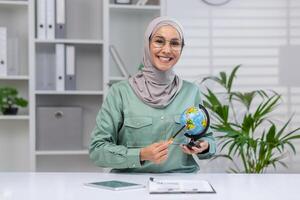 souriant femme portant une hijab, séance dans une brillant bureau, en portant une globe et montrer du doigt à il pour une leçon. concept de éducation, géographie, et diverse apprentissage. photo