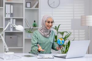 de bonne humeur femme dans hijab en portant globe pendant un en ligne appel dans une moderne bureau. concept de éducation, éloigné travail, et multiculturel communication. photo