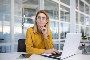 réfléchi femme d'affaires portant des lunettes et Jaune chemise travail sur une portable dans une moderne Bureau environnement. photo