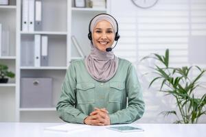 une de bonne humeur femme dans une hijab portant une casque, travail dans une moderne bureau, démontre professionnalisme et client soutien. photo