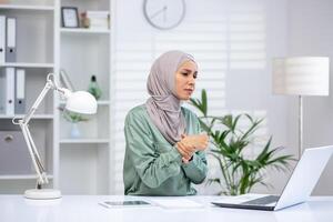 une concerné femme dans hijab en portant sa poignet dû à douleur tandis que travail sur une portable dans une brillant, moderne Bureau paramètre. photo
