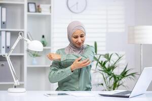 cette image capture une musulman femme dans une hijab expérience coude malaise tandis que travail à une bureau dans une brillant, contemporain bureau. photo