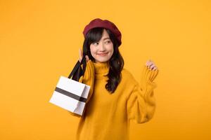 surpris Jeune asiatique femme dans sa 30s, en portant une achats papier sac, avec élégance dans une Jaune chandail et rouge béret contre une Jaune Contexte. photo