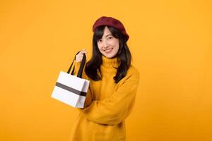 Jeune asiatique femme dans sa 30s, en portant une achats papier sac, rayonnant bonheur dans une élégant ensemble avec une Jaune chandail et rouge béret contre une Jaune Contexte. photo