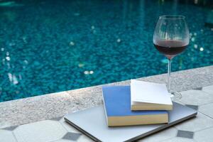 verre de rouge du vin et livres sur carnet près une nager bassin dans Matin de été. été Voyage concept. vacances et vacances concept. photo